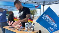 Parrakas nuorehko tumma ja tatuoitu mies asettelee tarroja, lippuja ja AfD-aiheisia koristeita telttakatoksen pöydän päälle.