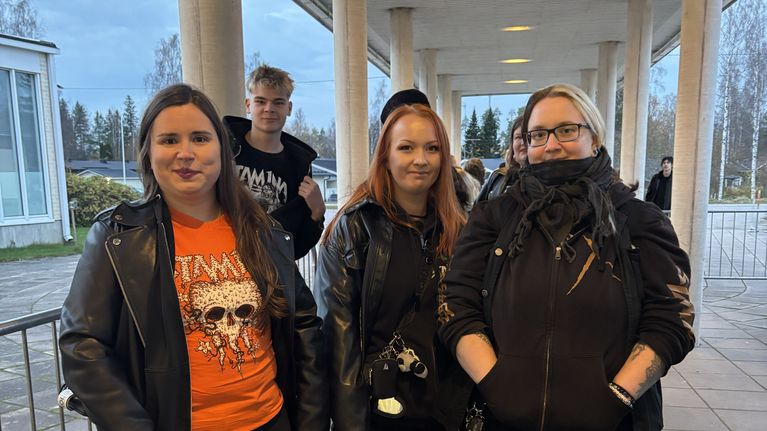 Viisi nuorta aikuista seisoo ulkona rakennuksen katoksen alla; edessä kolme henkilöä nahkatakeissa.