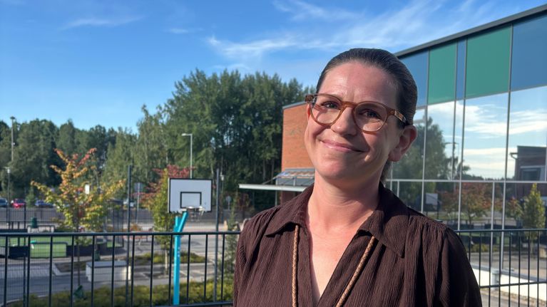 En person med glasögon står framför ett stängsel vid en basketplan utomhus.