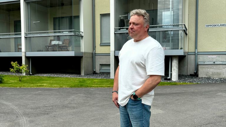 Valkoisen T-paitaan pukeutunut mies seisoo kerrostalon pihalla.