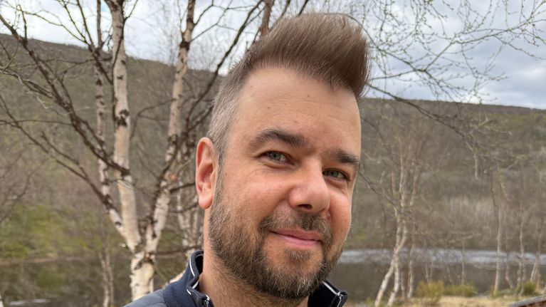 Ihminen seisoo keväisen jokimaiseman edessä. Hänellä on yllään kevyttoppatakki, kädet on taskuissa ja ilmeenä hymy.
