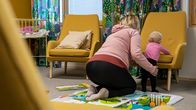 Alle vuoden ikäinen lapsi seisoo lattialla nojatuolin istuinta vasten. Hänen vieressään istuu lapsen äiti polvillaan ja pitää hellästi kiinni.