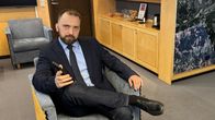 Mies istuu harmaalla nojatuolilla toimistohuoneessa, kädessään älypuhelin, taustalla televisio ja ikkuna.