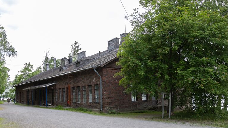 Tiilirakennus metsäisessä ympäristössä, oikealla puolella kyltti ja iso puu, edustalla soratie.