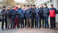 En grupp äldre män klädda för vinterpromenad och med gångstavar i händerna står uppställda på rad.