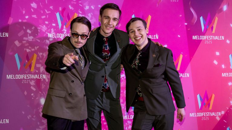 Axel Åhman, Kevin Holmström, och Jakob Norrgård poserar på röda mattan vid Melodifestivalen 2023. De är klädda i stiliga kostymer och står framför en bakgrund med festivalens logotyp.