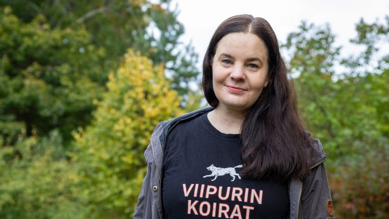 Aino Arjas seisoo ulkona ja katsoo kohti kameraa. Hänen yllään on paita, jossa lukee "Viipurin koirat".