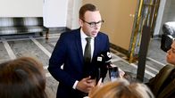 A man wearing glasses and a dark blue suit speaks to a crowd of reporters at the Parliament building. 