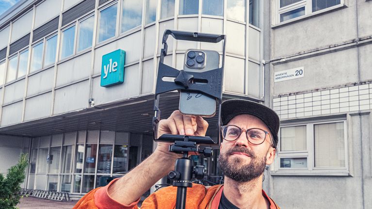 A photographer in front of a building with a camera, with the Yle logo on the building’s wall.