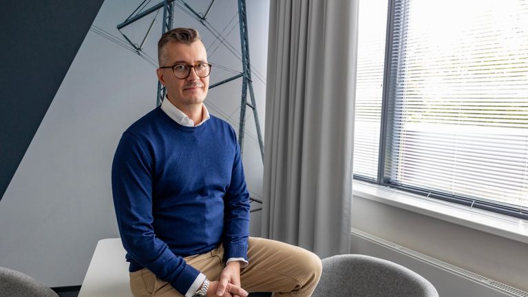 Keski-ikäinen mies, jolla on silmälasit ja sininen neulepaita, istuu kokoustilan pöydän reunalla ja katsoo kameraan.