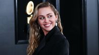 Miley Cyrusilla on pitkät hiukset kiharrettuna ja musta villakangastakki päällään. Hän katsoo sivulle olkansa yli ja hymyilee. 