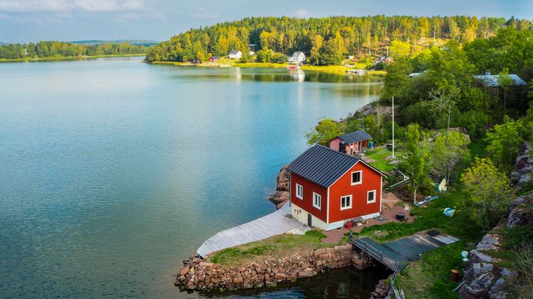 A red wooden building sits close to the shore in an idyllic summer landscape. 