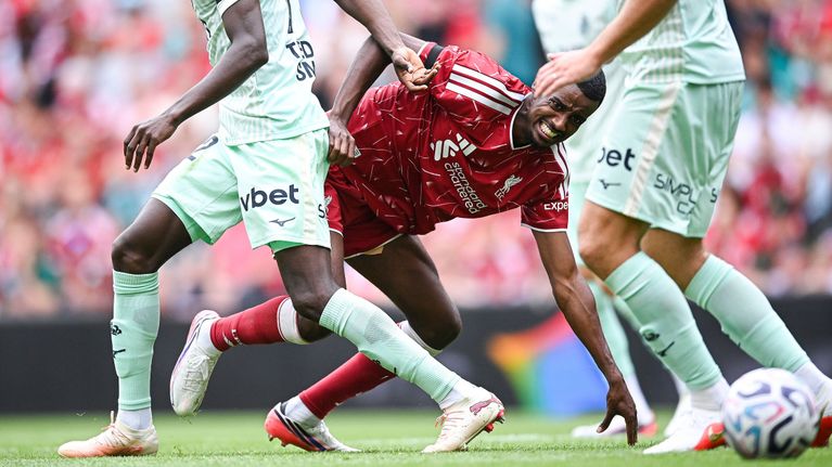 En fotbollsspelare i röd dräkt faller till marken mellan två motståndare i ljusgröna dräkter under en match.