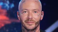 En man med blonderat hår och tatueringar på halsen småler. Han är klädd i vit t-skjorta och randig kavaj.