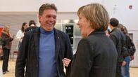 En leende person i polotröja och kavaj pratar med en annan i något som liknar en gymnastiksal. Längre bort i salen står andra människor.