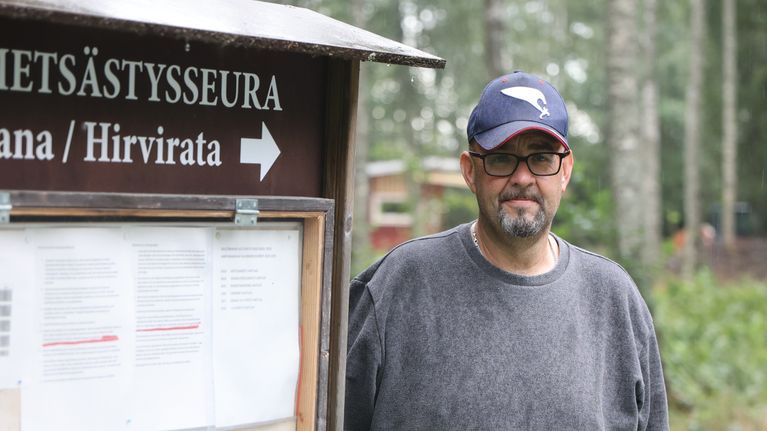 Dan Eriksson, en man med glasögon och keps står utomhus vid en anslagstavla med texten "METSÄSTYSSEURA", "ÄLGBANA/HIRVIRATA" och flera uppsatta papper.