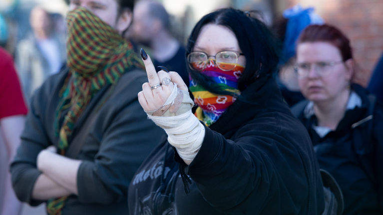Woman in black clothes with a rainbow-coloured cloth covering half of her face, gesturing with her hand and holding up her middle finger.
