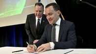 Kuvassa henkilö allekirjoittaa asiakirjan pöydän ääressä virallisessa tilaisuudessa.