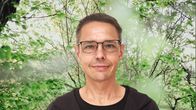 Olof Schybergson småler in i kameran. Bakgrunden är en grönskande skogsdunge.