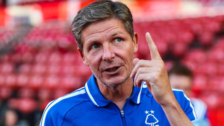 En man i blå träningsoverall med Nottingham Forests logotyp håller upp ett finger, suddiga läktare i bakgrunden.