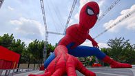Suuri Spider-Man-hahmopatsas kyykkyasennossa ulkoalueella, taustalla puita ja metallirakenteita.