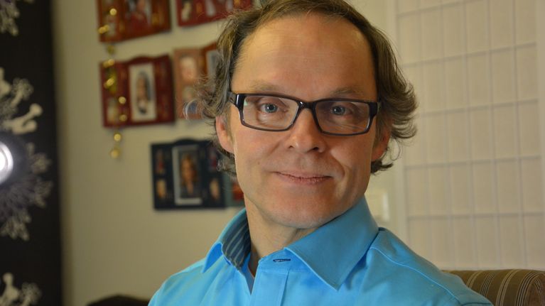 Ilkka Ilola katsoo kameraan, takana lasten muotokuvat seinällä