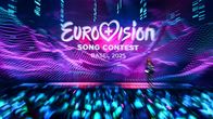 The Eurovision Song Contest 2025 logo on a screen behind a stage with a blurry announcer and audience.