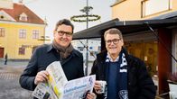 Kaksi miestä seisoo torilla. Toisella on käsissä Uutisrauma-lehti. Taustalla on keltainen vanha raatihuone.