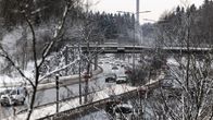 Talvinen kaupunkimaisema vilkkaan liikenneväylän varrella. Autoja kulkee pitkin moottoritietä lumen peittämien puiden ja rakennusten keskellä.