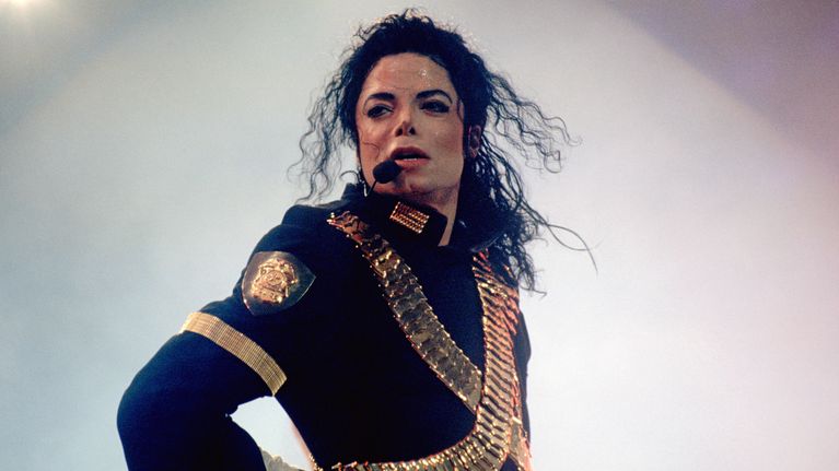 Michael Jackson katsoo ohi kamerasta sinertävässä konserttiasustuksessa, langaton mikrofoni suulla.