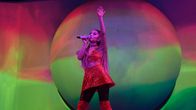 Laulaja Ariana Grande seisoo värikkään screenin edessä lavalla toinen käsi ilmaan nostettuna. Toinen käsi pitelee mikkiä suun edessä. Päällään hänellä on punaiset korkeat saappaat ja punainen paljettiasu.