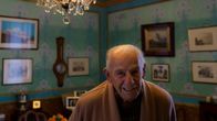 98-vuotias Antonio Grippo kotonaan huoneessa, jossa turkoosit seinät ja kattokruunu sekä seinillä paljon vanhoja valokuvia.