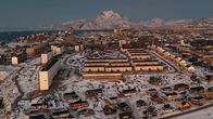 Flygfoto på en liten stad med villor, radhus och flervåningshus i ett snöigt landskap vid havet, snöklädda berg i bakgrunden.