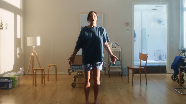 En kvinna klädd som patient står framför en sjukhussäng i sin lägenhet. Hon håller ut händerna.  