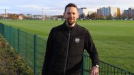 En svartklädd man lutar sig mot ett staket bredvid en fotbollsplan.