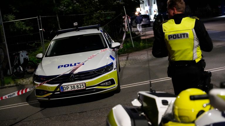 Tanskalainen moottoripyöräpoliisi selkä kameraan päin, ja poliisiauto kadulla iltapimeällä.