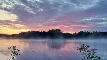 Järvimaisema auringonlaskun aikaan, veden pinnalla näkyy sumua ja taustalla metsäinen ranta.