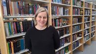 En leende kvinna med blont hår i pagefrisyr framför bokhyllor på ett bibliotek.