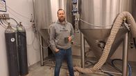 Skäggprydd man i grå tröja med kaffekopp i handen framför stort kärl i rostfritt stål.