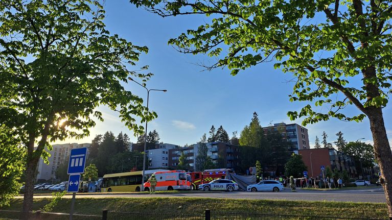 An ambulance, a police car and a bus are parked on a street next to apartment buildings, with people gathered nearby.
