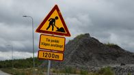 Varningsskylt vid väg med texten "Tie poikki Vägen avstängd 1200m", stenhög och grå himmel i bakgrunden.