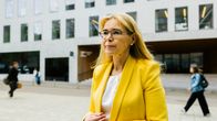 En kvinna med långt blont hår och gul kavaj står utomhus framför en modern byggnad med många fönster.