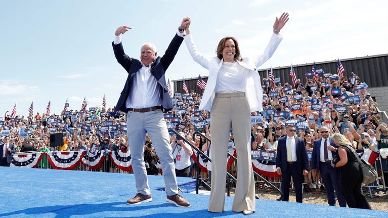 Tim Walz ja Kamala Harris seisovat vierekkäin lavalla kädet ylhäällä pitäen toisiaan kädestä kiinni. Taustalla yleisöä Yhdysvaltain lippujen ja kampanjakylttien kanssa.