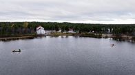Kaksi venekuntaa soutaa järvellä, taustalla rannalla valkoisia rakennuksia ja metsäistä maisemaa.