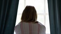 A person sits on a bed looking out of a window.
