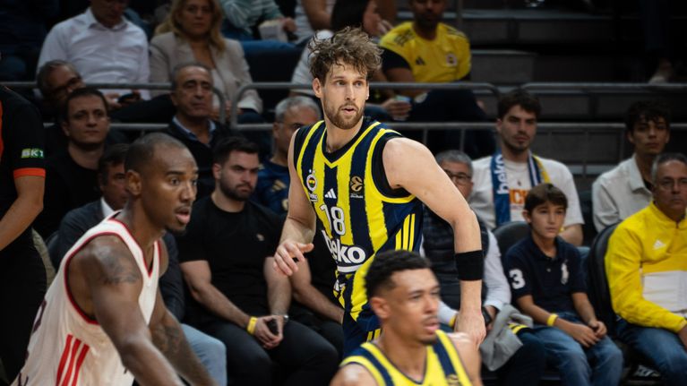 Kuvassa on koripallo-ottelu, jossa pelaajat Fenerbahçen peliasussa ja punavalkoisessa peliasussa kilpailevat kentällä.
