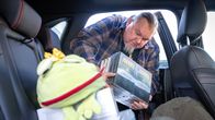 Esikoiskirjailija ottaa kirjoja auton takapenkiltä, etualalla sammakkopehmolelu.