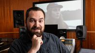 En leende man med skägg sitter i en studio. En stor videoskärm bakom honom.