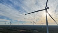 Eight wind turbines sticking out of a forest, seen against a partly cloudy sky from the air.