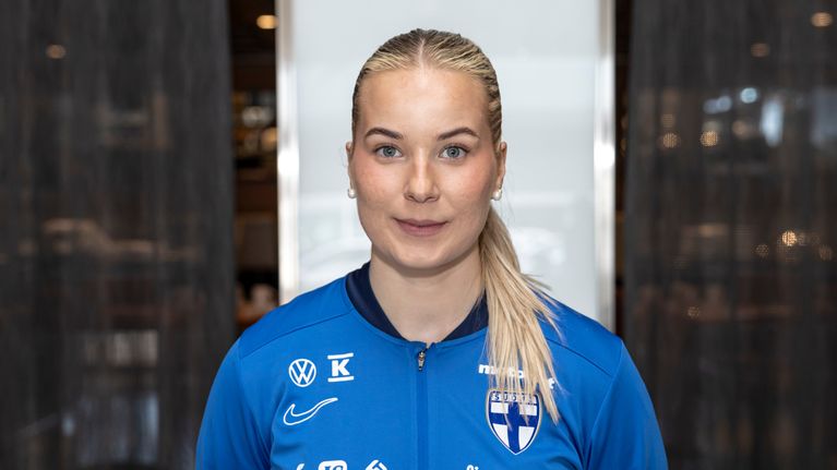 Nainen sinisessä jalkapallo-paidassa katsoo kameraan ja hymyilee. Hänellä on pitkät hiukset.